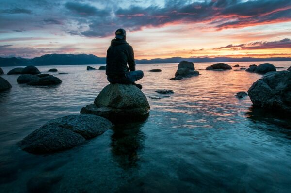 Person meditiert auf einem Felsen am Wasser während des Sonnenuntergangs – Meditation auf Reisen