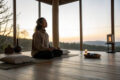 Person praktiziert Stille Meditation in einem ruhigen, minimalistischen Raum mit Sonnenaufgang im Hintergrund.